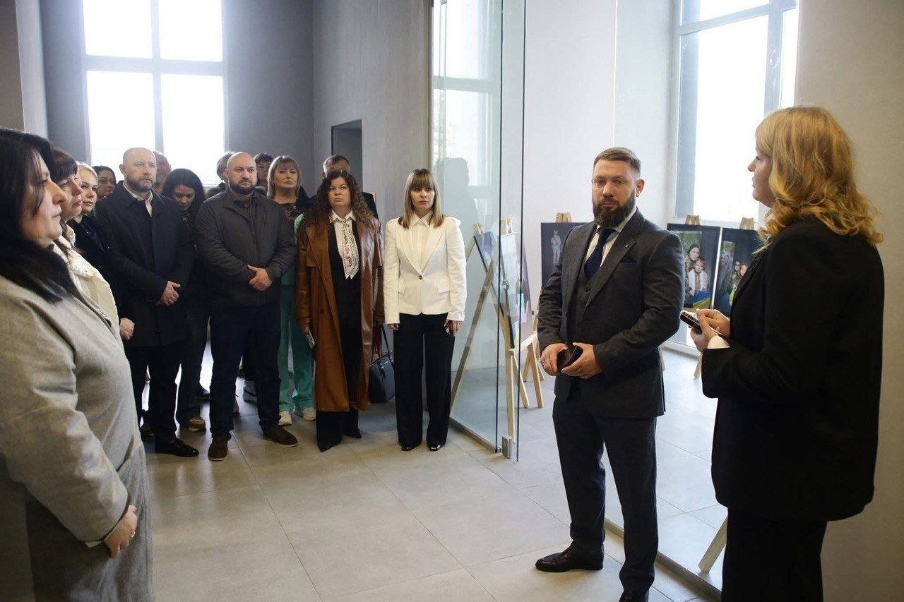 Група людей у діловому одязі зібралася у світлому сучасному приміщенні. Чоловік та жінка на передньому плані звертаються до присутніх, а на фоні, за скляною перегородкою, видно художню виставку з картинами на мольбертах.