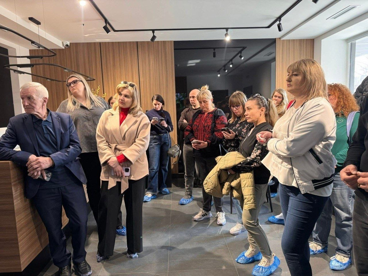 Група людей у бахілах стоїть у сучасному світлому приміщенні з дерев'яним оздобленням. Присутні уважно слухають, деякі з них тримають у руках смартфони.