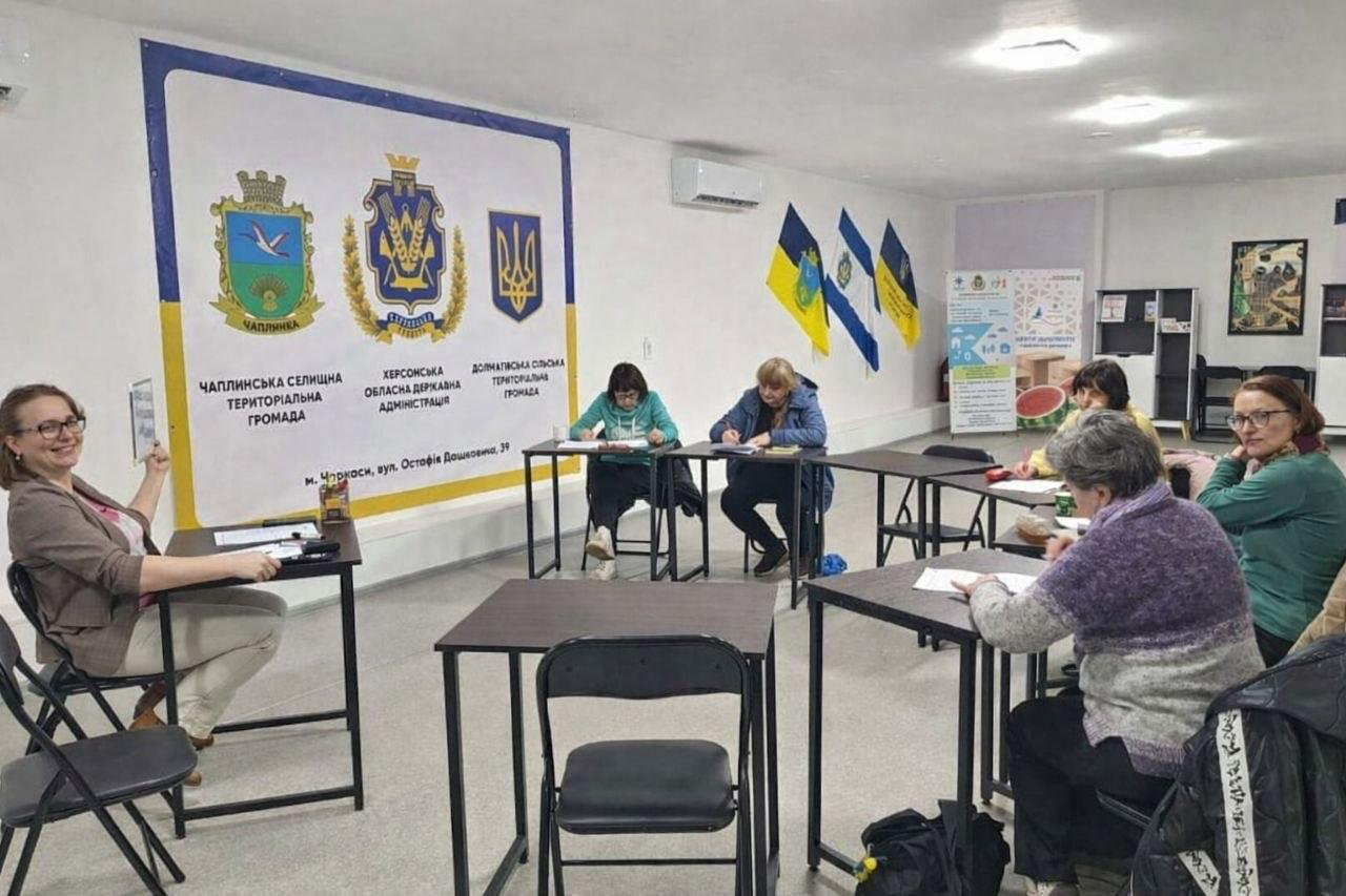 Група людей працює за столами в аудиторії під час навчального заходу чи робочої зустрічі. На стіні розміщено великий банер із гербами та назвами Чаплинської, Долматівської громад і Херсонської ОДА, а також встановлено прапори.