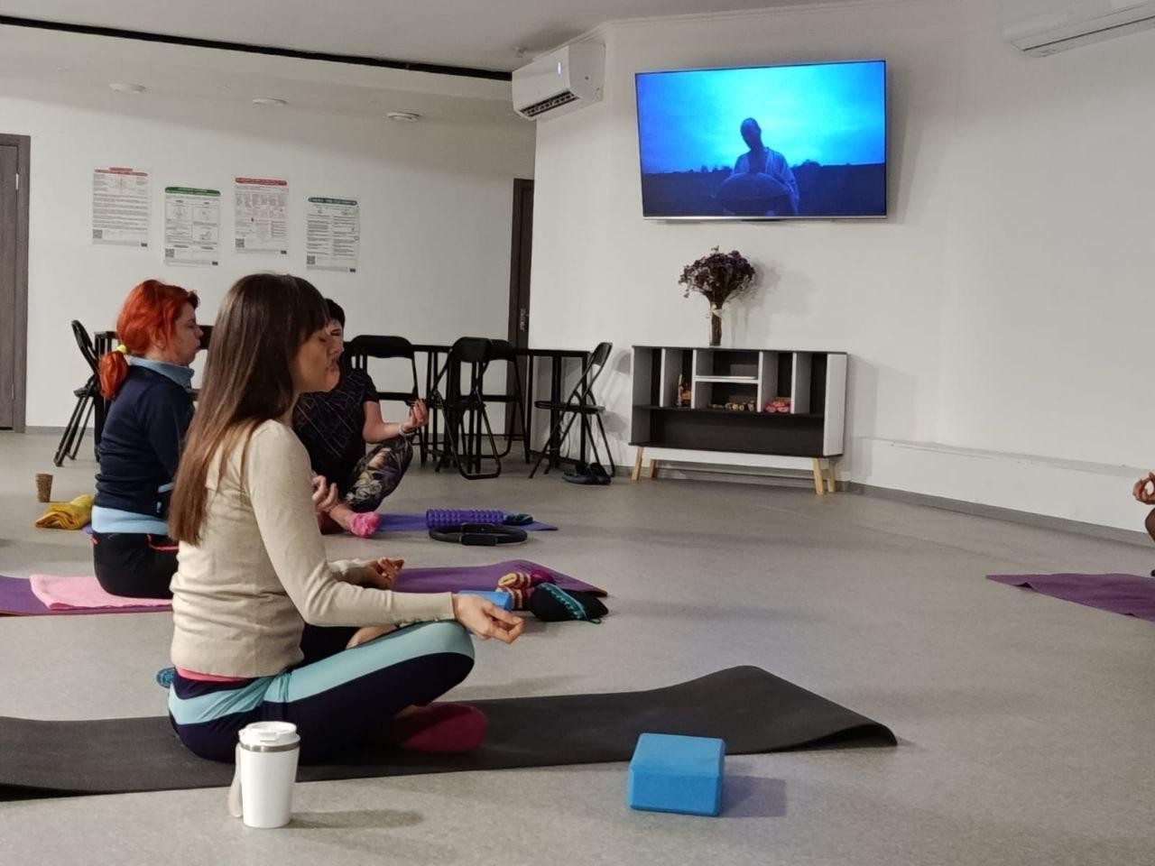 Група жінок займається йогою або медитацією у світлому залі, сидячи на килимках у позі лотоса. На стіні перед ними висить телевізор, на якому транслюється відео.