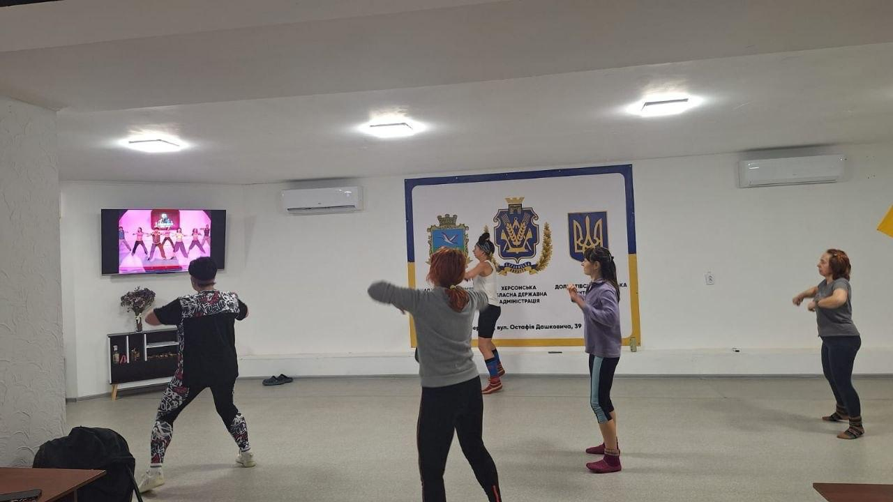 Група жінок займається фітнесом або танцями у світлому залі, повторюючи рухи за відео на настінному екрані. На стіні розміщено великий банер із символікою Херсонської обласної державної адміністрації.