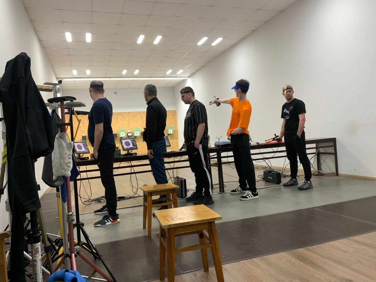 Група чоловіків у спортивному одязі тренується у стрілецькому тирі. Вони стоять у ряд перед мішенями, один із них (у яскравій помаранчевій футболці) зафіксований у момент прицілювання з пневматичного пістолета у витягнутій руці.