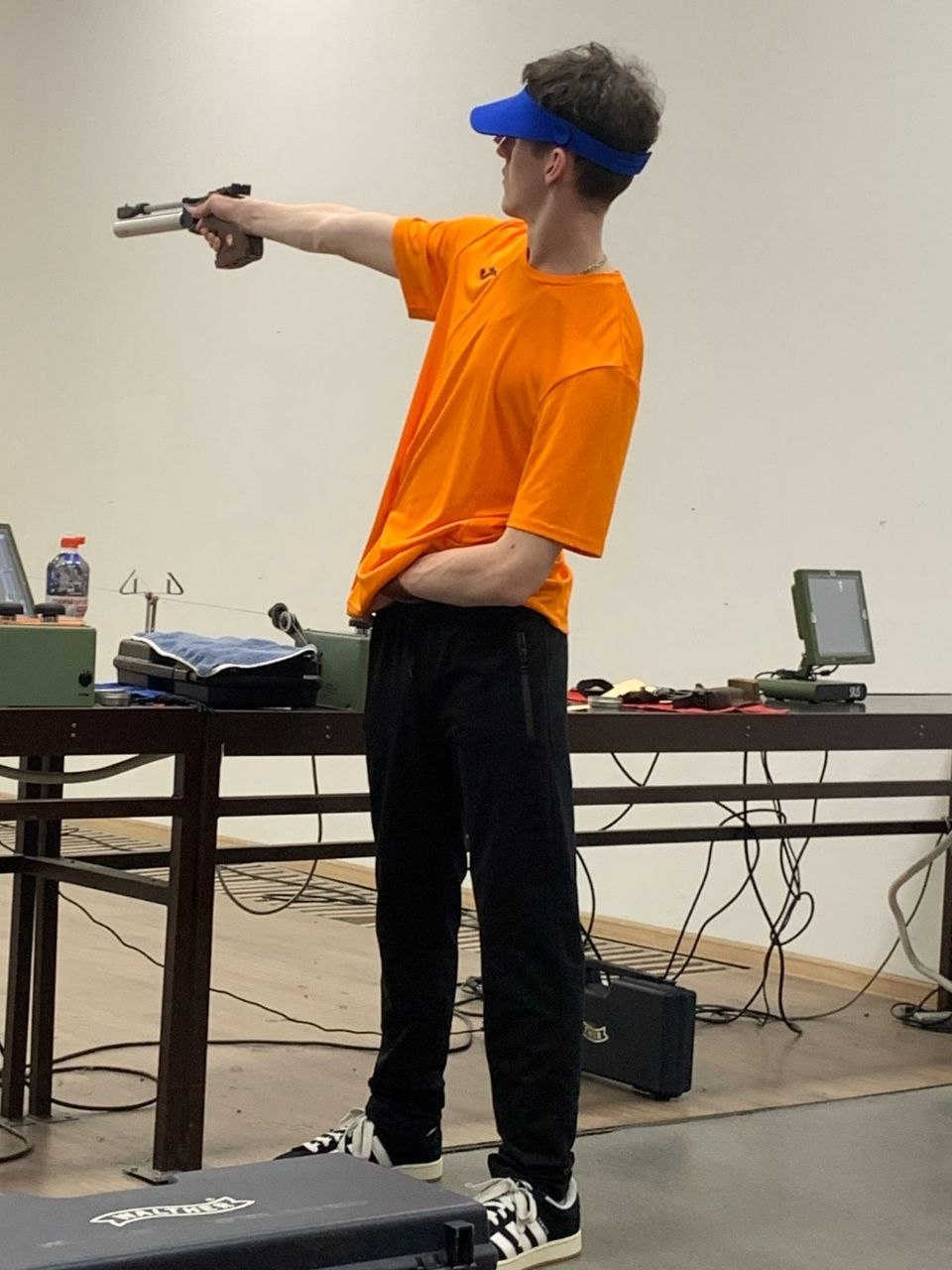 Молодий чоловік у спортивному одязі (помаранчева футболка, чорні штани та синій козирок) займається спортивною стрільбою. Він стоїть у профіль, тримаючи пневматичний пістолет у витягнутій руці, цілячись у мішень у приміщенні тиру.