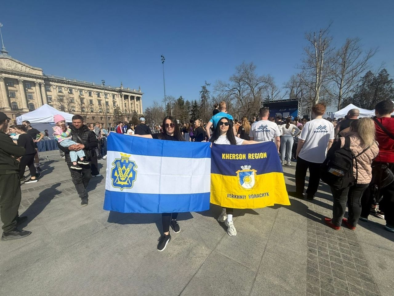 Дві дівчини тримають прапори Херсонщини та селища Верхній Рогачик на міській площі під час масового заходу. На задньому плані видно натовп людей, намети та адміністративну будівлю під ясним небом.