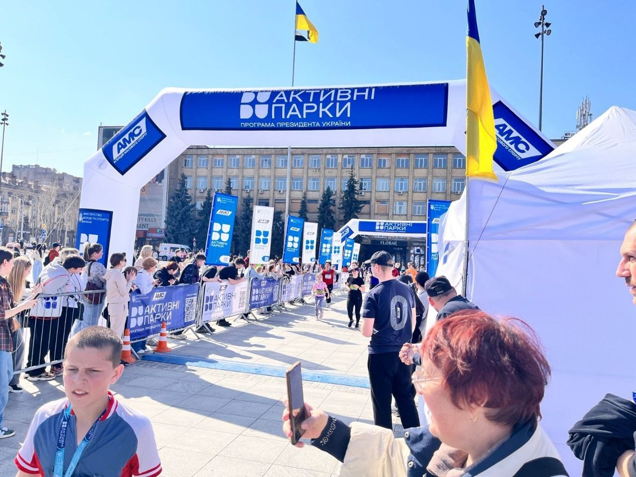 Масовий спортивний захід на міській площі в межах програми «Активні парки». На фоні адміністративної будівлі встановлено брендовану надувну арку, прапори України та огороджену трасу, вздовж якої зібралися глядачі та учасники.