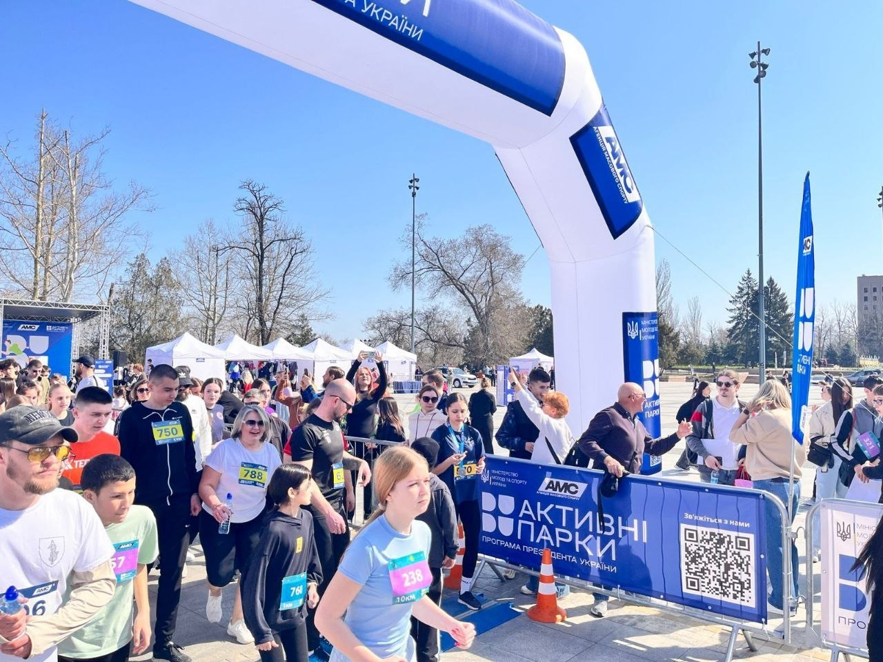 Масовий спортивний забіг у межах програми «Активні парки». Учасники з номерами на одязі проходять під великою біло-синьою надувною аркою на міській площі в сонячний день.