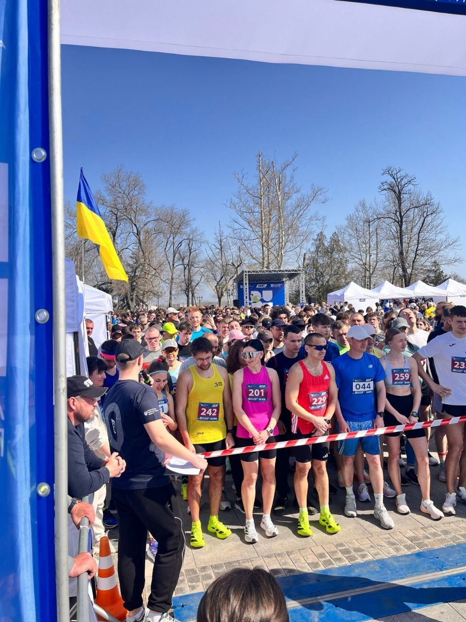 Великий натовп учасників забігу на лінії старту під відкритим небом у сонячний день. Бігуни в яскравому спортивному одязі з номерами чекають сигналу за обмежувальною стрічкою, на фоні видно прапор України та сцену.