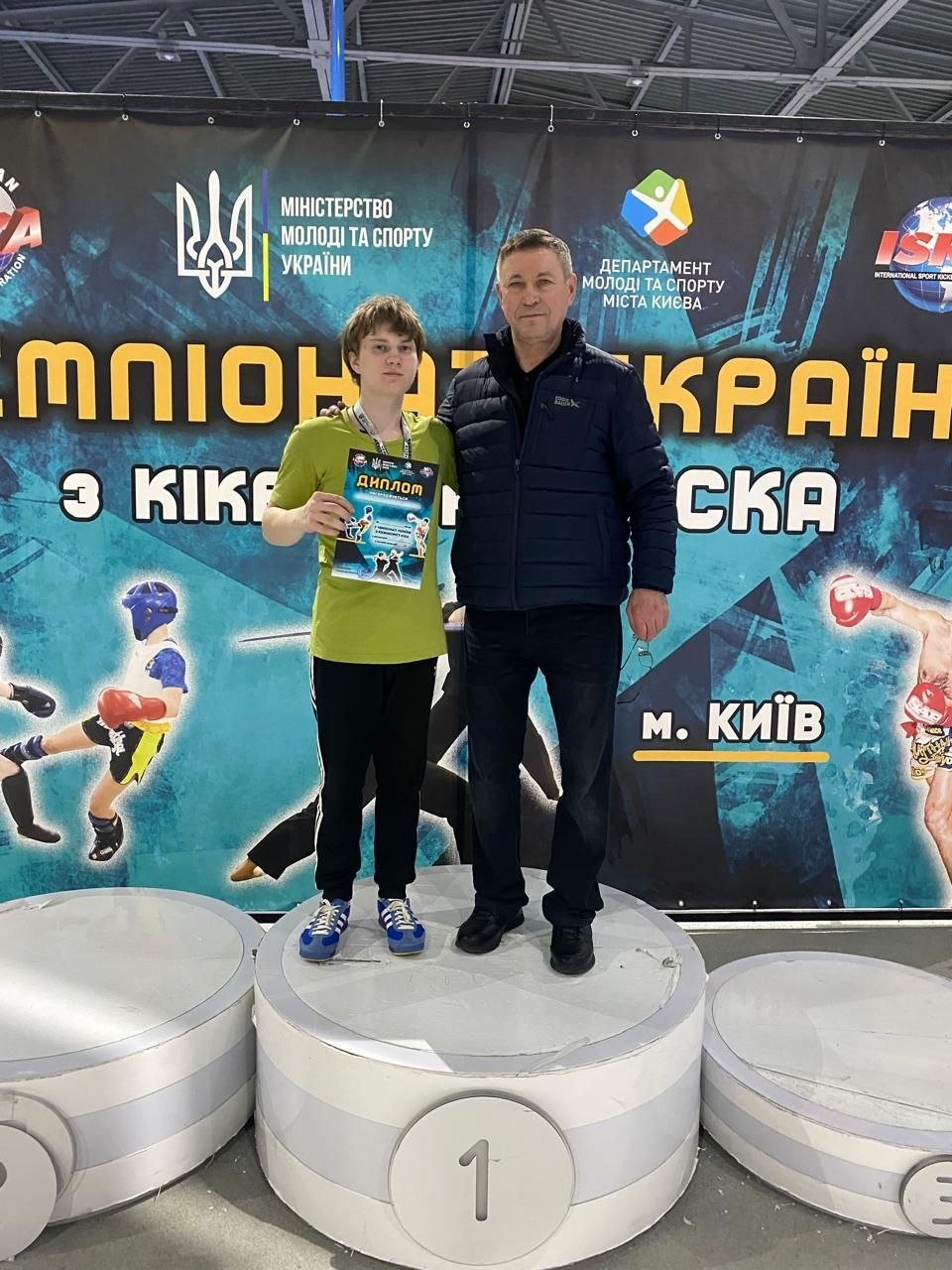 Нагородження переможця на Чемпіонаті України з кікбоксингу ISKA у Києві. Молодий спортсмен із медаллю та дипломом стоїть на першій сходинці п'єдесталу поруч із чоловіком у спортивній куртці на фоні офіційного банера змагань.