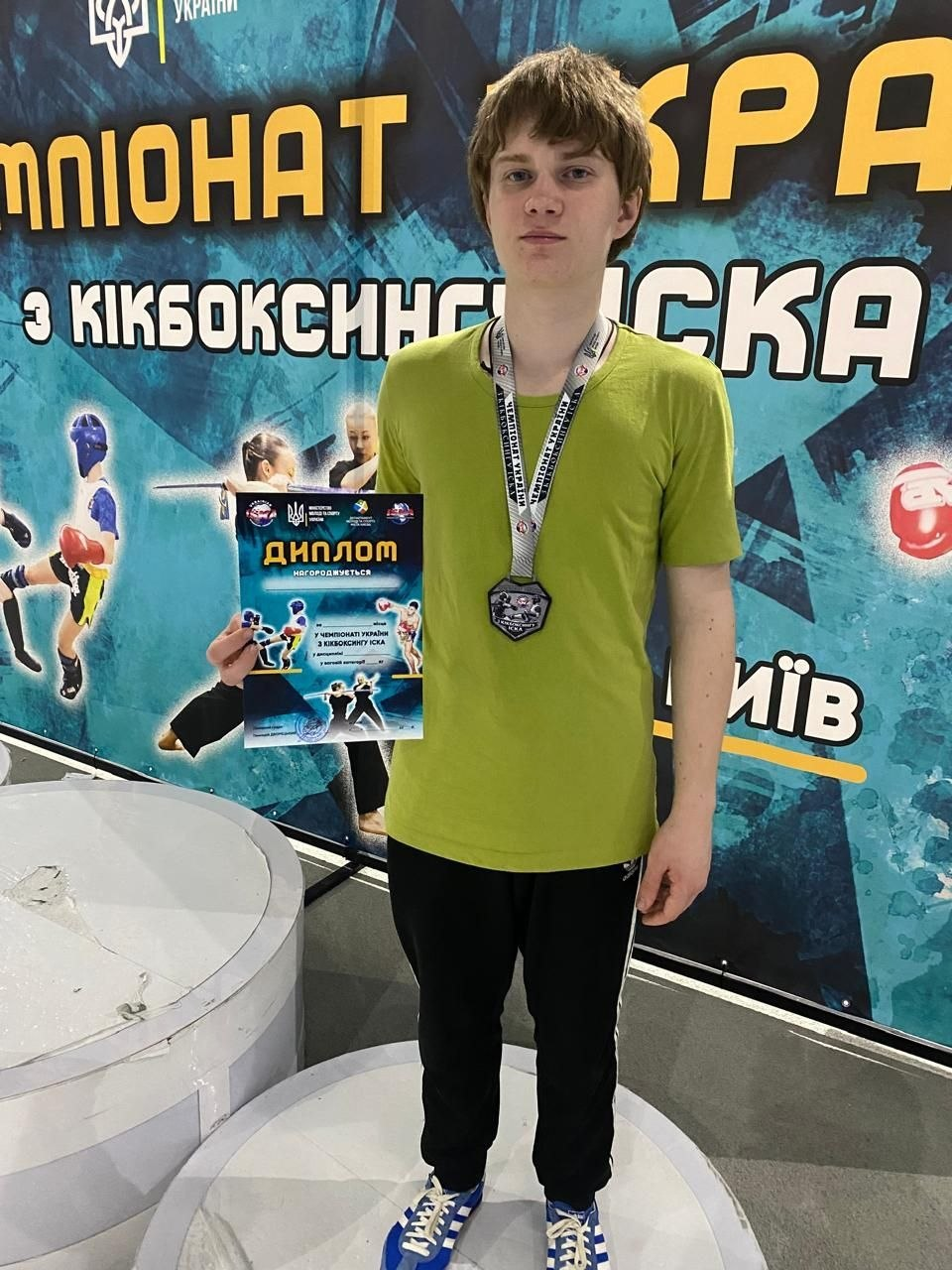 Молодий чоловік стоїть на п'єдесталі на фоні банера «Чемпіонат України з кікбоксингу ISKA». Він одягнений у салатову футболку та спортивні штани, тримає в руці диплом, а на шиї у нього медаль.