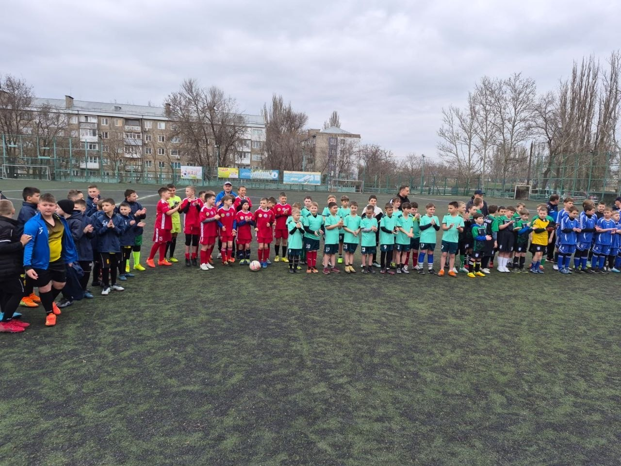 На футбольному полі зібралося кілька дитячих команд у різноколірній формі (червоній, бірюзовій, синій та чорній). Юні спортсмени вишикувалися в ряд на фоні житлових будинків та дерев під час проведення змагань або тренування.