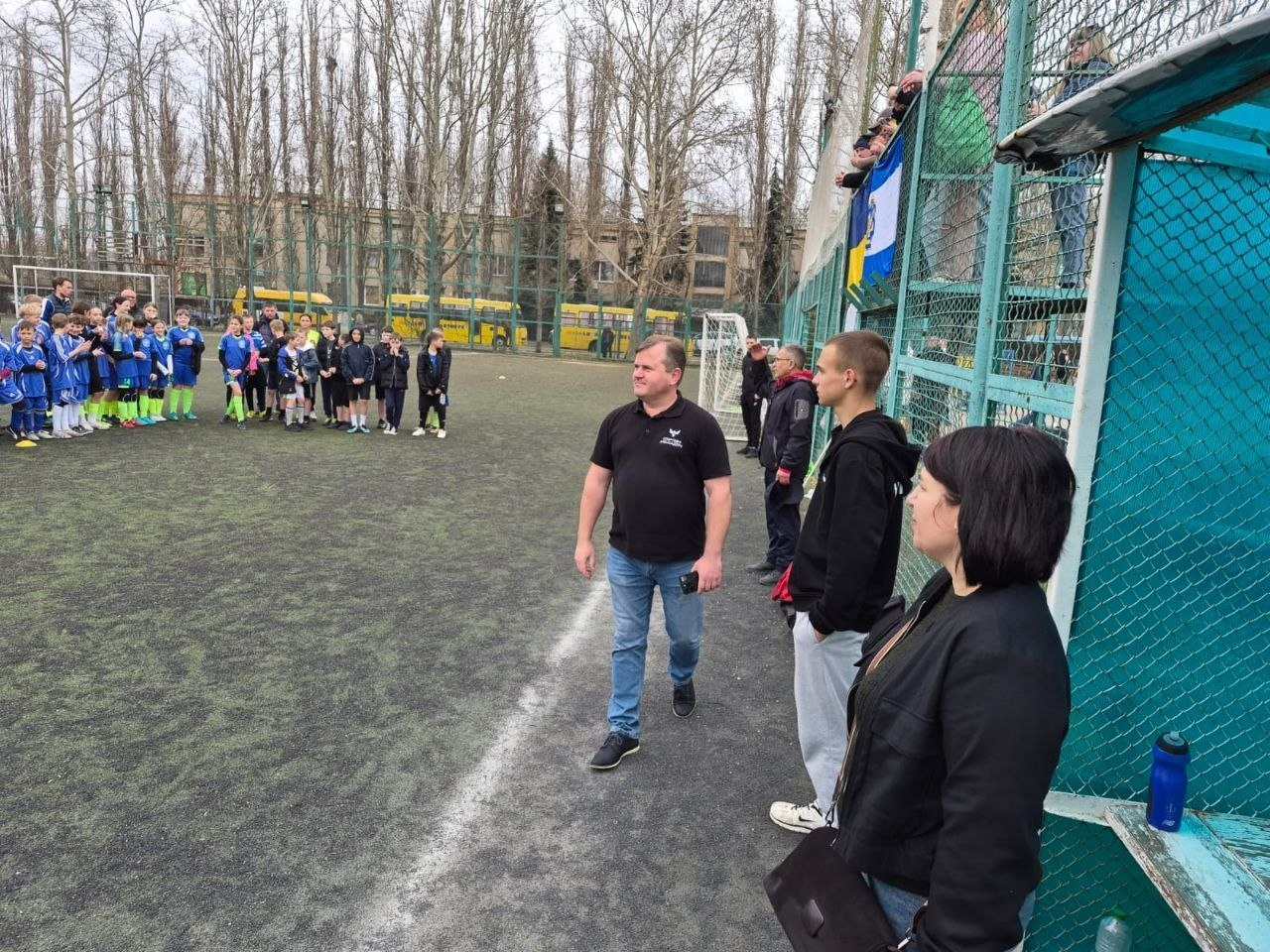 Футбольне поле зі штучним покриттям, на якому група дорослих спостерігає за дітьми у спортивній формі, що вишикувалися в ряд. На задньому плані за сіткою паркана видно високі дерева без листя, жовті автобуси та глядачів.