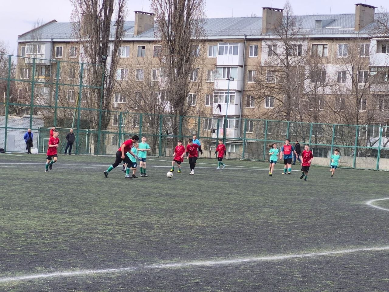 Молоді футболісти у червоній та бірюзовій формі грають у футбол на огородженому майданчику зі штучним покриттям. На фоні видно багатоповерховий житловий будинок та дерева.