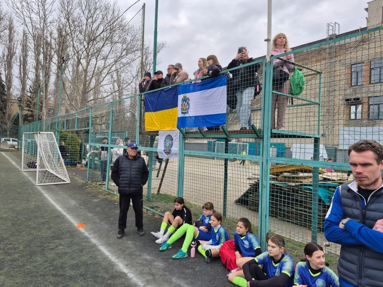Група юних футболісток у синьо-зеленій формі відпочиває на краю футбольного поля зі штучним покриттям. Над ними на трибунах знаходяться вболівальники з прапором України та спортивною символікою, а поруч стоять тренери або організатори заходу.