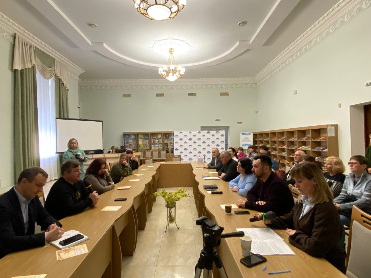 У залі засідань за довгим П-подібним столом проходить зустріч або конференція. Група чоловіків та жінок у повсякденному та діловому одязі обговорюють робочі питання; на столах лежать документи, стоять паперові стаканчики та мікрофони.