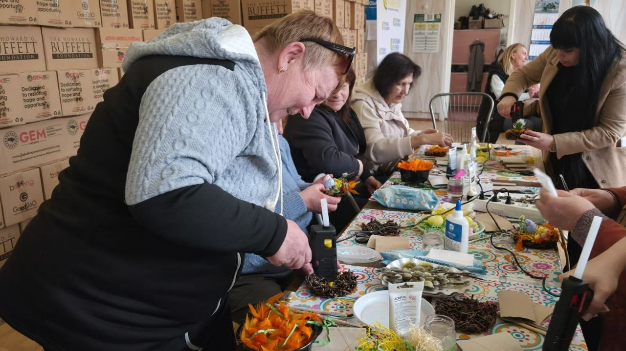 Майстер-клас із декорування: група жінок за довгим столом створює святкові композиції, використовуючи клейові пістолети, штучні квіти та природні матеріали. На задньому плані складено коробки з гуманітарною допомогою від міжнародних фондів.