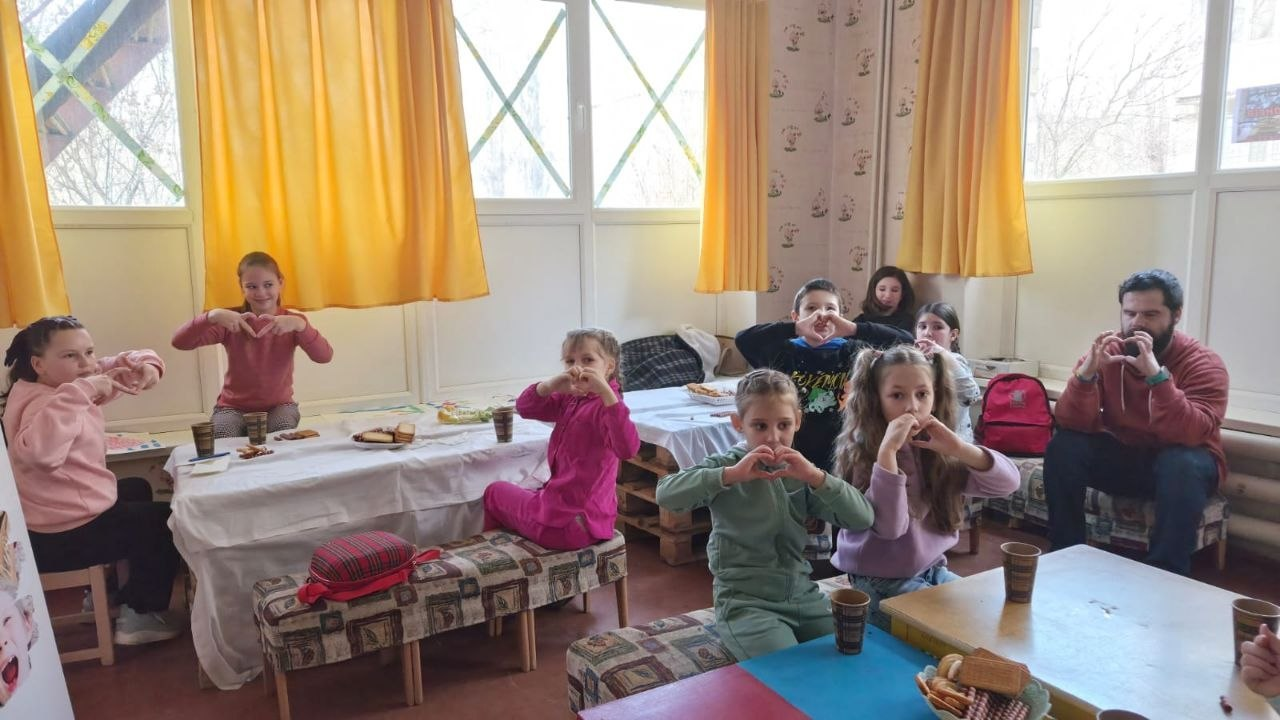 Група дітей та дорослих сидять за столами у світлій кімнаті з жовтими шторами та разом показують жест «серце» руками. На столах стоять стаканчики з напоями та тарілки з печивом.