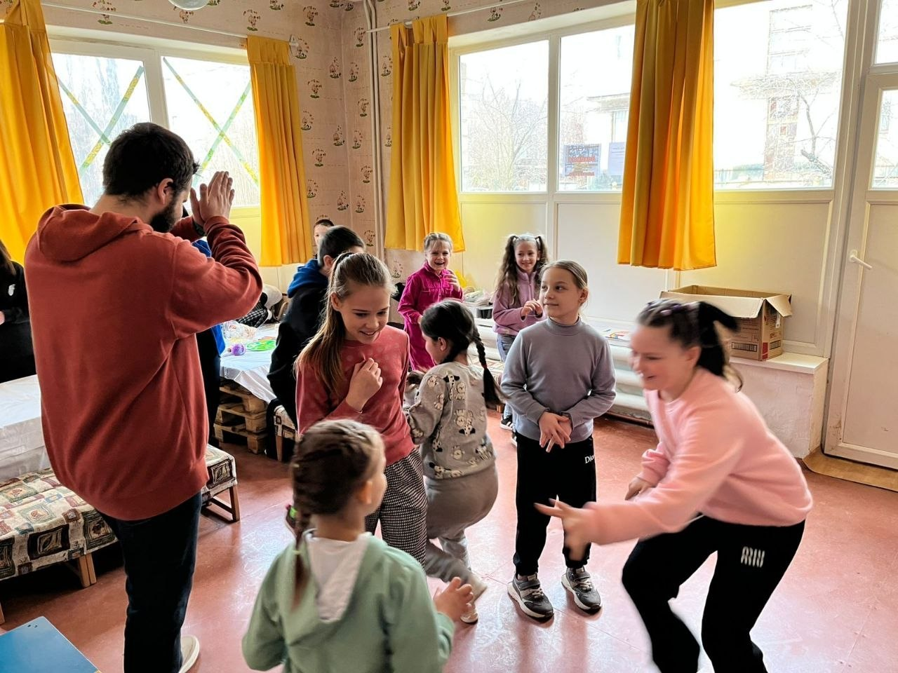 Група дітей активно грається та розважається у світлій кімнаті з великими вікнами та жовтими шторами. Чоловік у червоному худі показує рухи або дає вказівки під час спільної гри чи тренування.