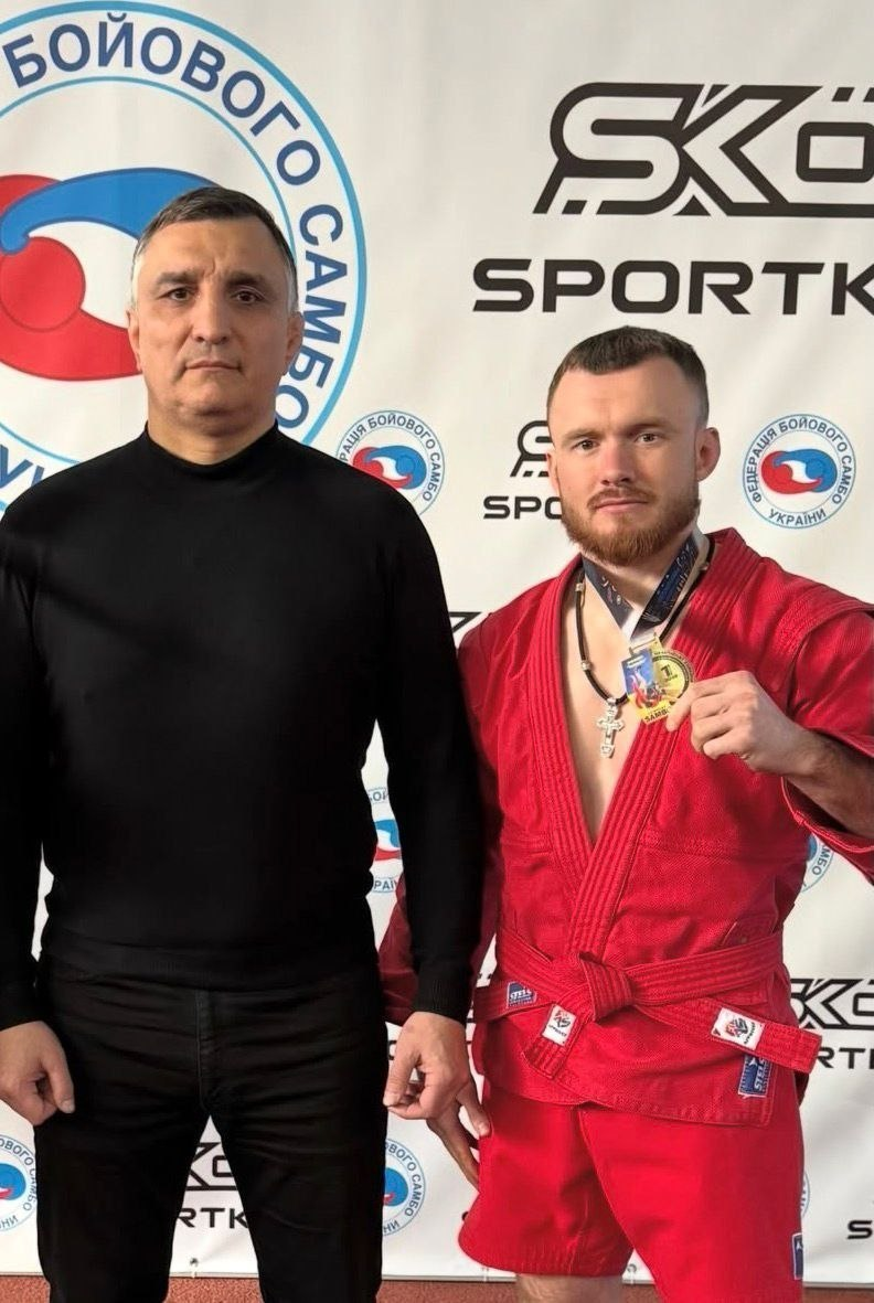 Двоє чоловіків на фоні банера Федерації бойового самбо України. Один — у чорному гольфі та штанах, інший — у червоному куртці-кімоно (самбовці) з золотою медаллю на шиї, яку він тримає рукою.