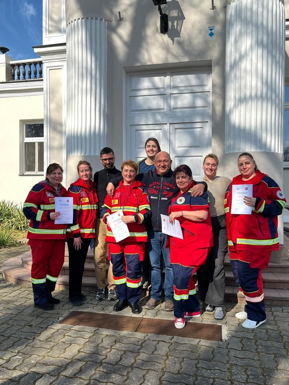 Групова фотографія людей на фоні будівлі з колонами. Частина присутніх одягнена в уніформу Товариства Червоного Хреста та тримає в руках сертифікати.