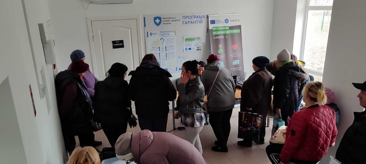 Група людей у черзі в приміщенні медичного або соціального закладу. На стінах розміщені інформаційні стенди «Програма медичних гарантій» та банер організації «Casers».