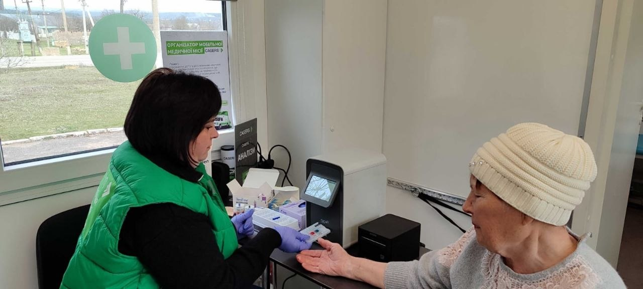 Медичний працівник у зеленому жилеті бере кров з пальця у літньої жінки для експрес-аналізу в мобільному медичному пункті.