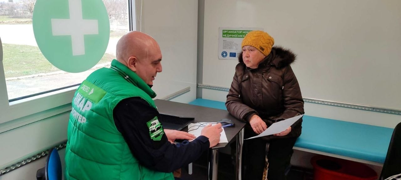 Лікар у зеленому жилеті консультує жінку в мобільному медичному пункті. На вікні розміщена наклейка із зеленим медичним хрестом.
