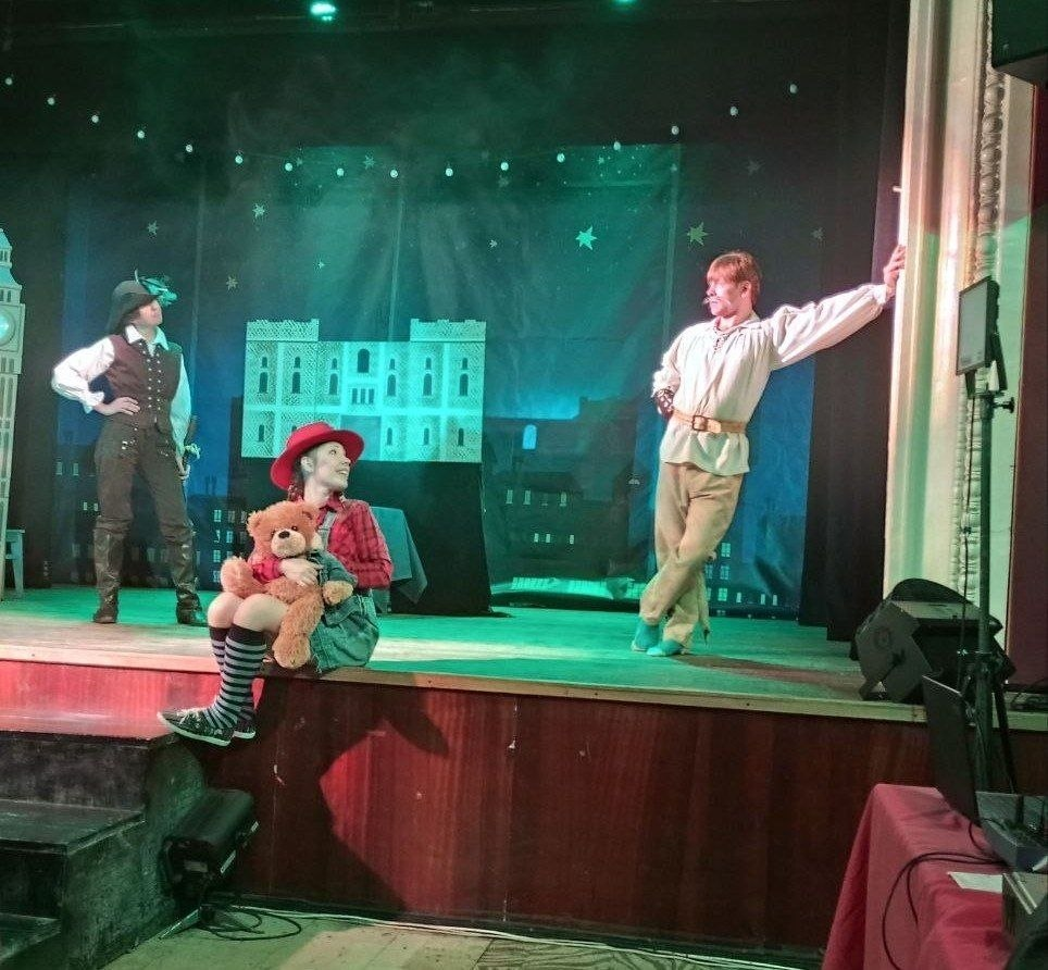 Театральна вистава: троє акторів у казкових костюмах на сцені з декораціями нічного міста. На передньому плані сидить дівчина з плюшевим ведмедем.