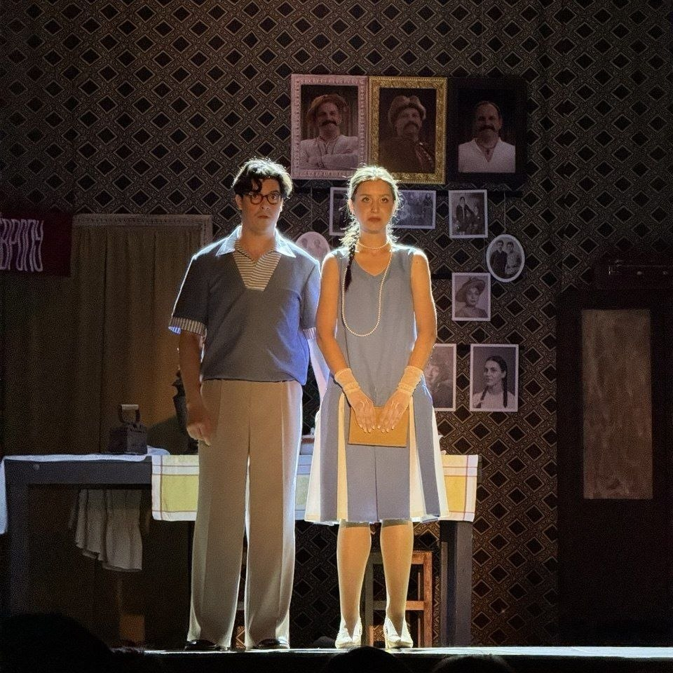 Театральна сцена: чоловік і жінка в ретро-вбранні стоять перед стіною з фотографіями в рамках.