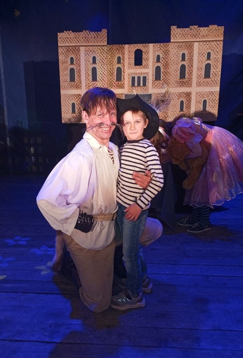 На сцені стоять чоловік у театральному костюмі Кота в чоботях та хлопчик у смугастій кофті. На задньому плані — декорація замку.