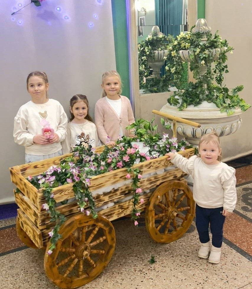 Четверо дітей позують біля декоративного дерев'яного воза, прикрашеного квітами, у світлому приміщенні з фонтаном на задньому плані.