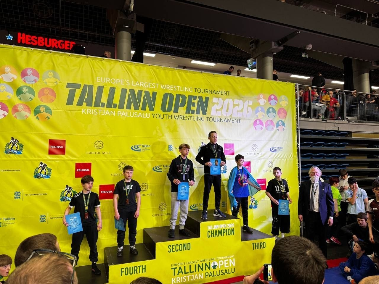Церемонія нагородження юних борців на п'єдесталі під час турніру Tallinn Open 2026. Переможці тримають грамоти на фоні великого жовтого банера зі спонсорами.