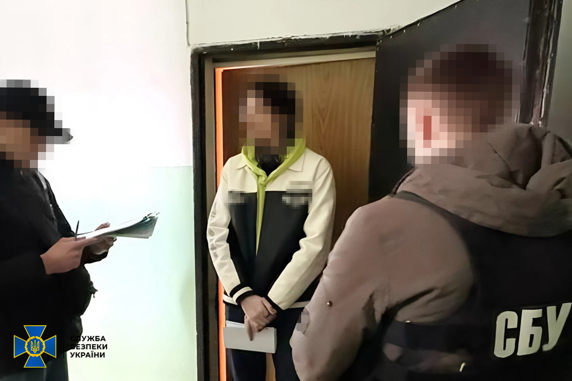 Співробітники СБУ проводять процесуальні дії у дверях приміщення. На передньому плані — представник служби у бронежилеті з написом «СБУ», ліворуч людина занотовує інформацію, а в дверному отворі стоїть затримана особа; обличчя всіх присутніх затушовані.