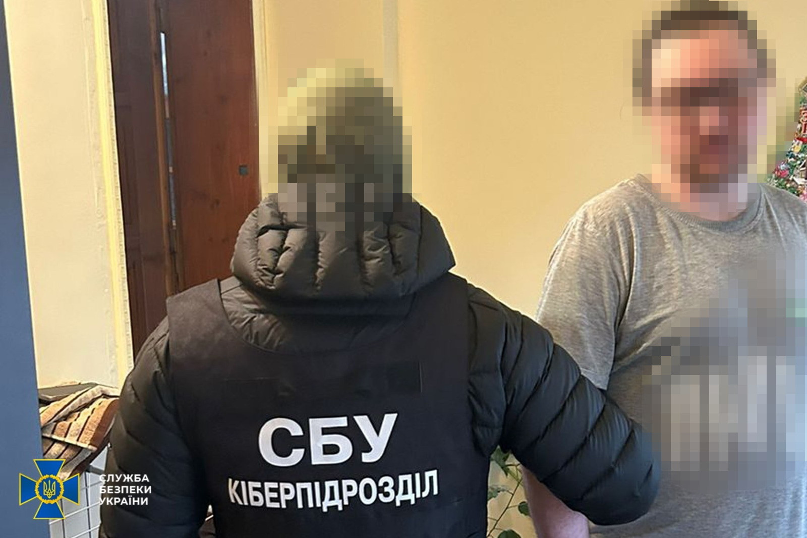 Представник кіберпідрозділу СБУ у форменому жилеті стоїть спиною до камери поруч із чоловіком у цивільному одязі; обличчя обох осіб затушовані.
