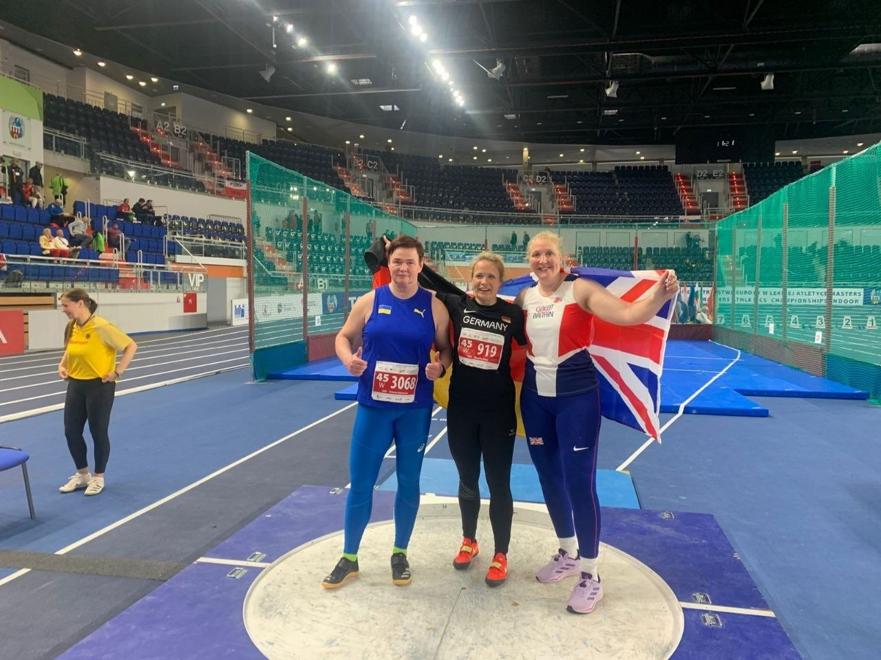 Три спортсменки в легкоатлетичній формі збірних України, Німеччини та Великої Британії позують у секторі для штовхання ядра на критих спортивних змаганнях.