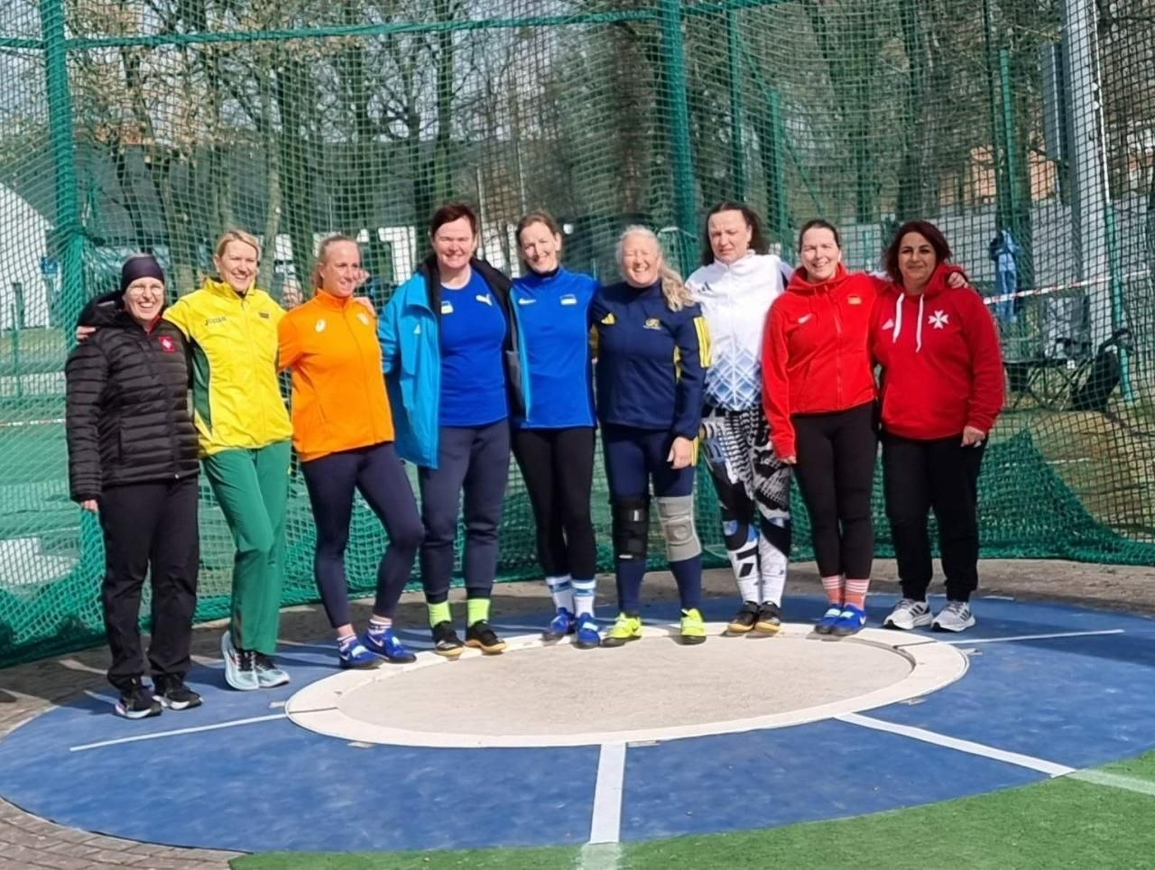 Група жінок-спортсменок у різноколірній спортивній формі позує для колективного фото в секторі для метань на відкритому стадіоні на фоні захисної сітки.