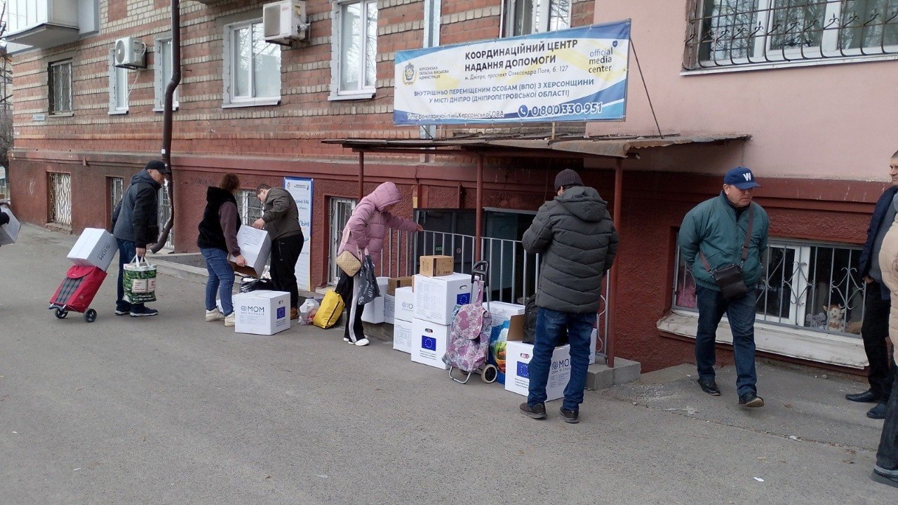 Група людей отримує гуманітарну допомогу біля входу до координаційного центру. На вулиці стоять картонні коробки з маркуванням міжнародних організацій, а над входом розміщено банер «Координаційний центр надання допомоги».