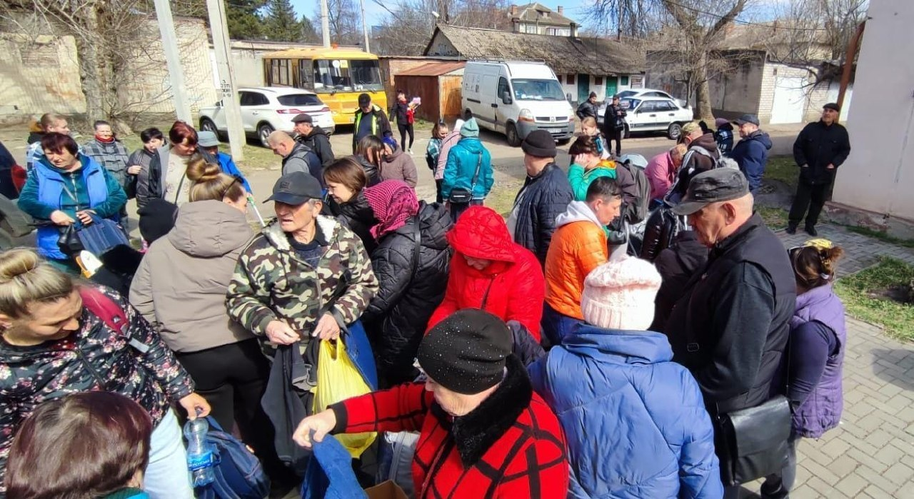 Група людей різного віку в повсякденному одязі зібралася на вулиці в житловому секторі. На задньому плані видно припарковані автомобілі, мікроавтобус та жовтий автобус.