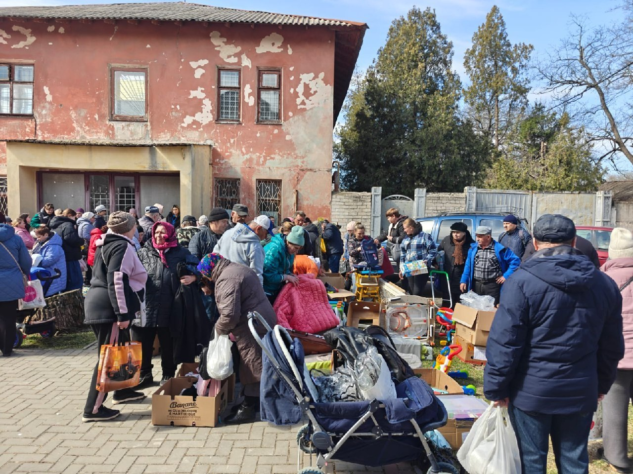 Група людей на вулиці біля старої будівлі отримує гуманітарну допомогу. На передньому плані — дитячий візочок, картонні коробки та пакети з речами.
