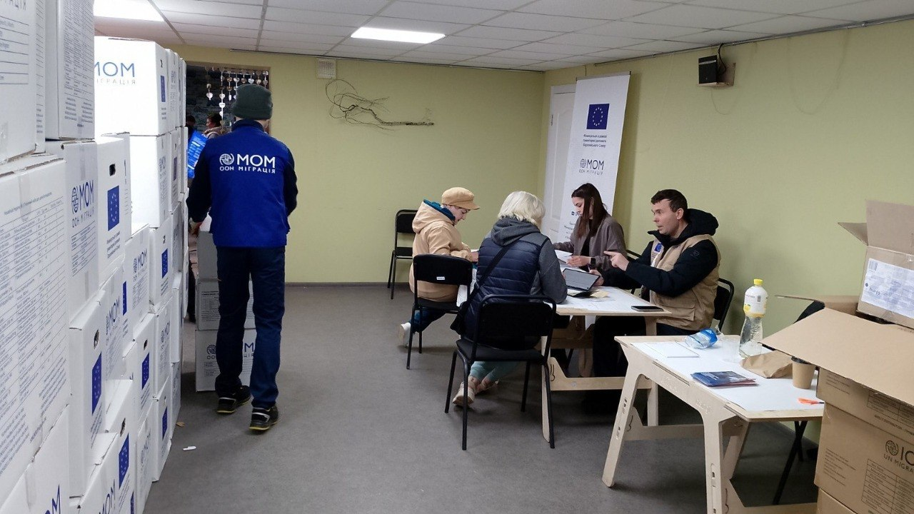 Пункт реєстрації для отримання допомоги в приміщенні. Співробітники в жилетах за столами спілкуються з відвідувачами, а поруч складено високі стоси коробок із гуманітарною допомогою з логотипами міжнародних організацій.