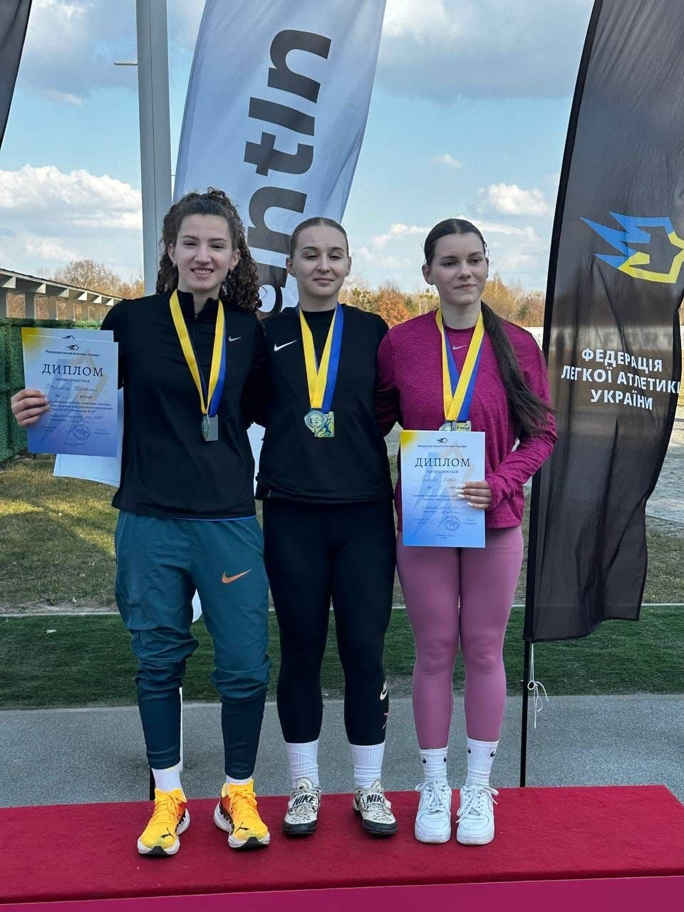 Три спортсменки стоять на п'єдесталі пошани з медалями на грудях та дипломами в руках. Позаду них видно прапор з написом «Федерація легкої атлетики України».