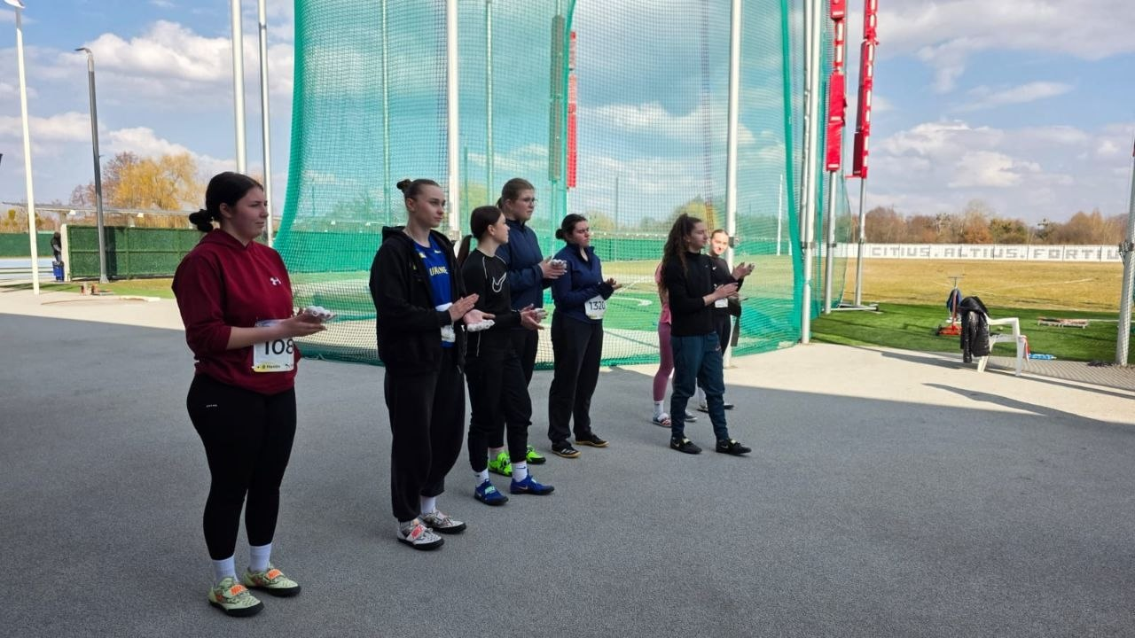 Група спортсменок у спортивній формі стоїть на майданчику для легкоатлетичних метань. Позаду них видно високу захисну сітку та олімпійське гасло «Citius, Altius, Fortius» на огорожі.