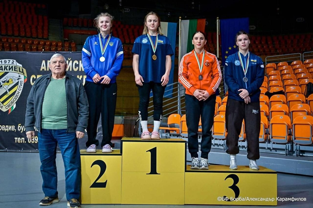 Чотири спортсменки з медалями на шиї стоять на призовому подіумі в спортивному залі. Поруч із ними, зліва від п'єдесталу, стоїть чоловік у повсякденному одязі. На задньому плані видно порожні глядацькі трибуни з помаранчевими сидіннями та прапори різних країн.