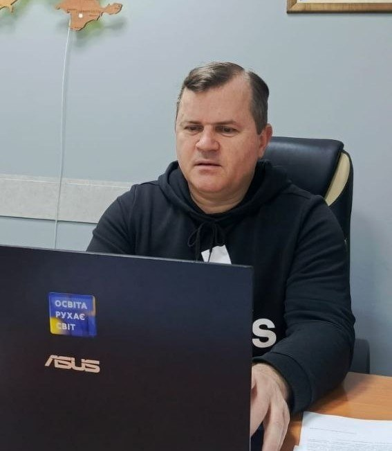 Чоловік у чорному худі працює за ноутбуком у робочому кабінеті. На кришці ноутбука наклейка з написом «Освіта рухає світ», а на стіні позаду видно частину дерев’яної мапи України.