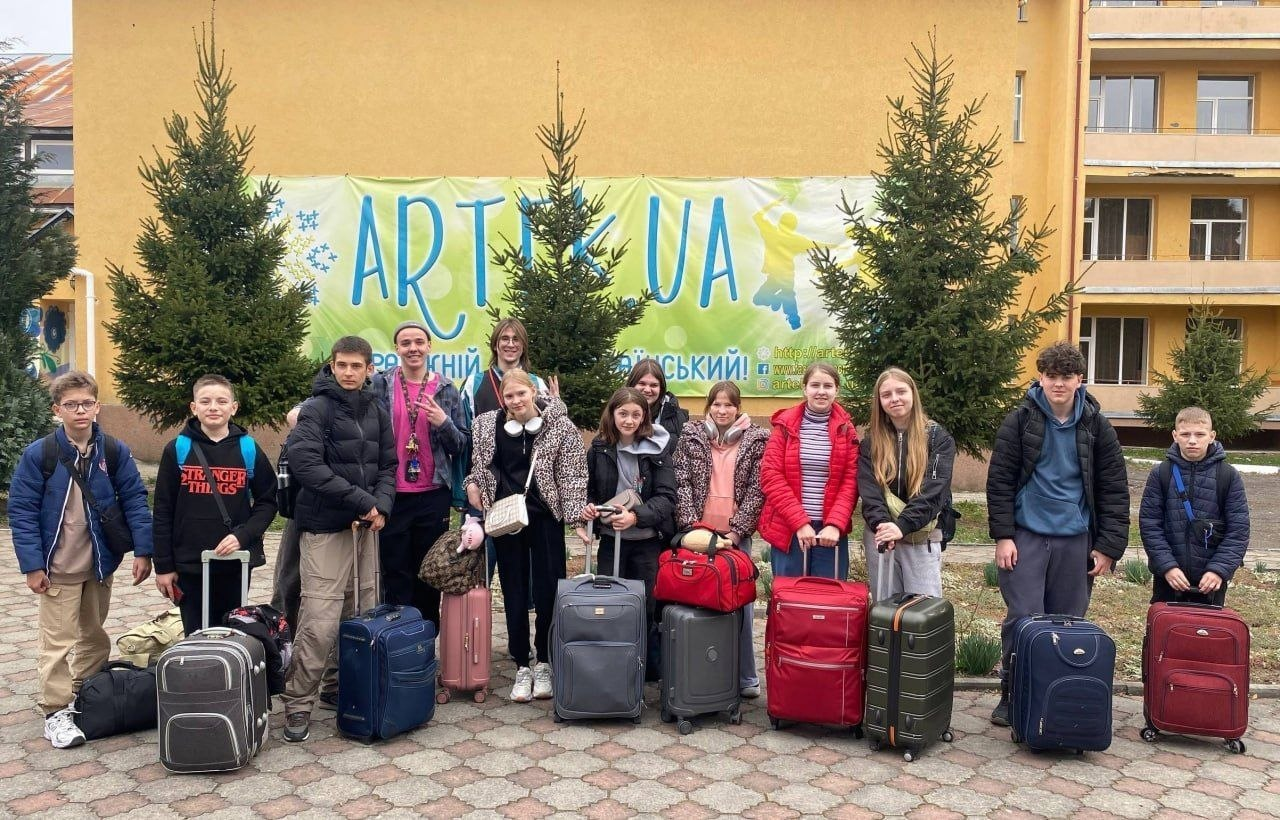 Група підлітків із валізами та сумками стоїть на подвір'ї будівлі. Позаду них розміщено банер із написом «ART UA» та зображенням ялинкових гілок.