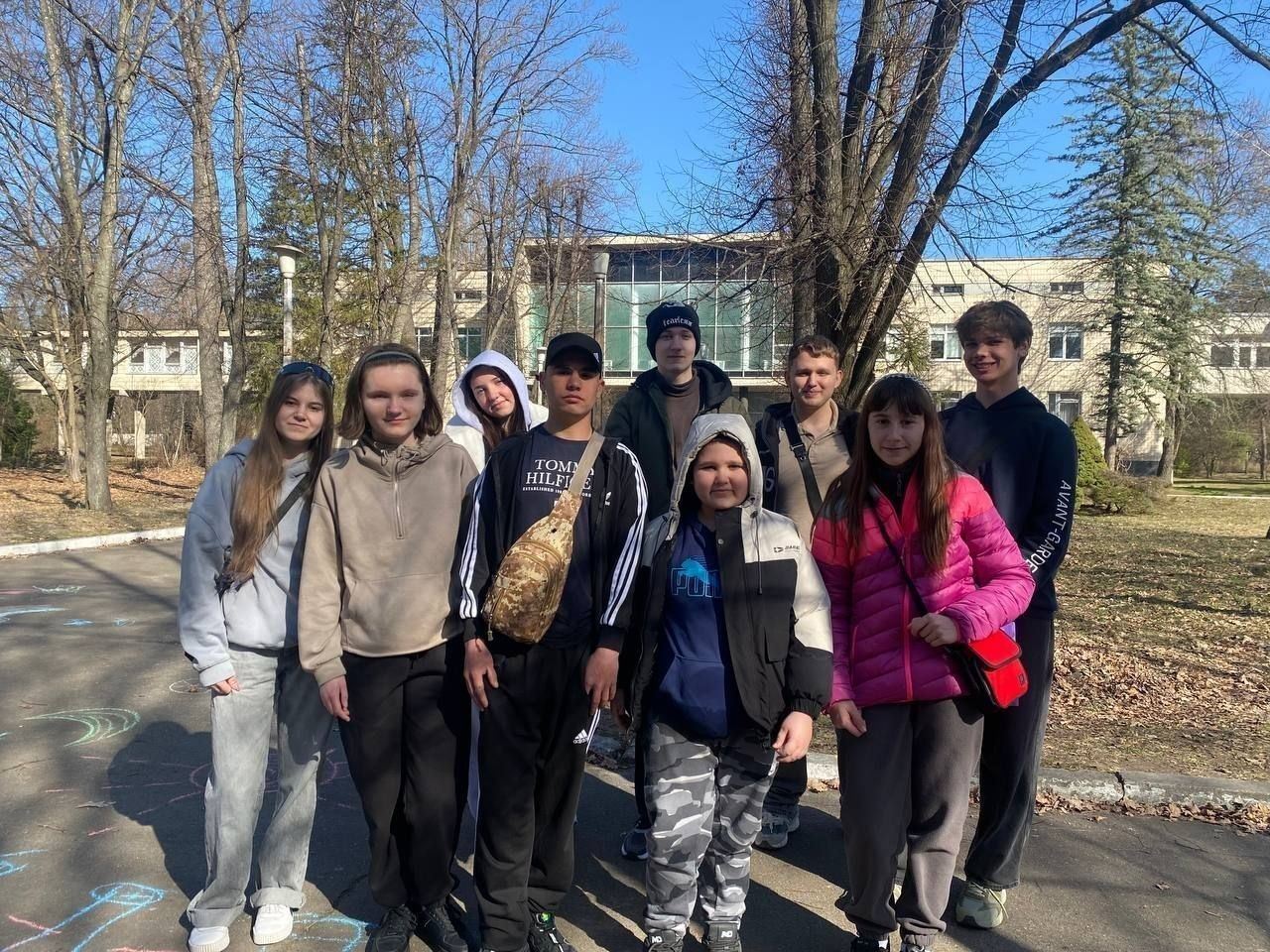 Група підлітків у весняному одязі позує для фото на асфальтованій доріжці в парку або на подвір'ї закладу. На задньому плані видно дерева без листя та світлу двоповерхову будівлю з великими вікнами.
