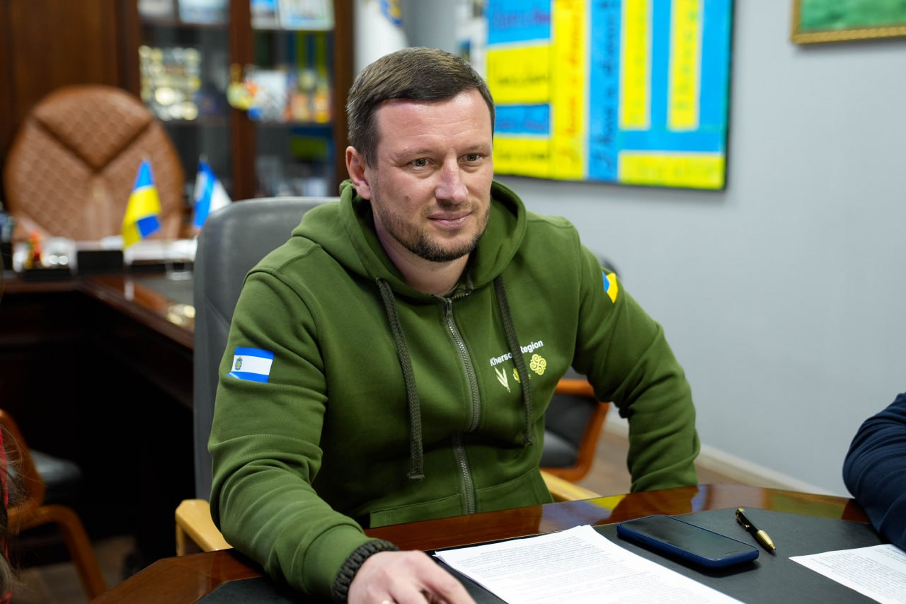 Чоловік у зеленому худі з українською символікою сидить за робочим столом у кабінеті. Перед ним лежать документи та смартфон, а на фоні видно офісні меблі та прапор України.