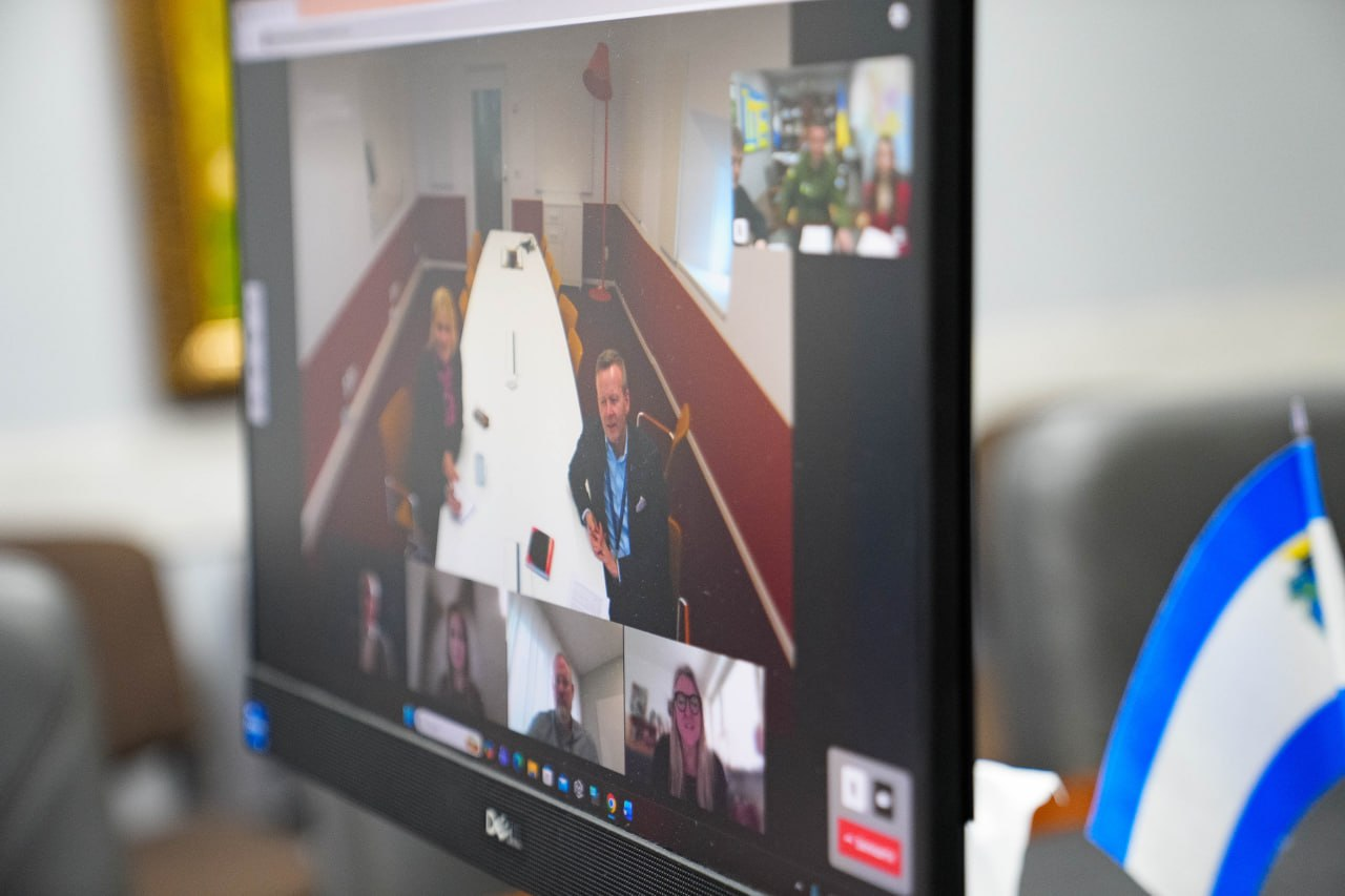 На екрані монітора триває відеоконференція за участю декількох осіб у різних вікнах. На передньому плані поруч із монітором стоїть настільний прапорець, а на фоні видно розмиті деталі кабінету.