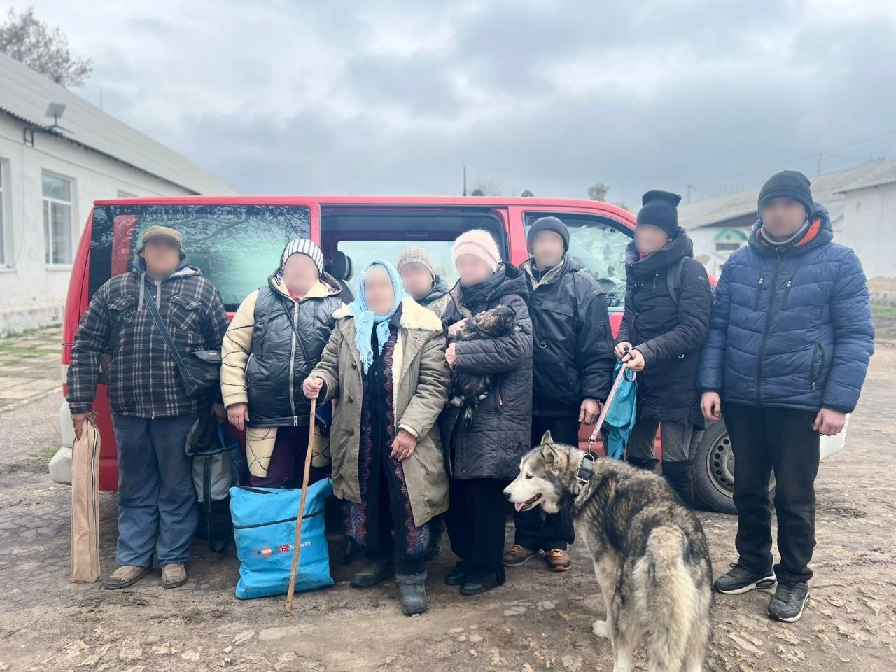 Група з восьми людей у теплому одязі з особистими речами та собакою породи хаскі стоїть перед червоним мікроавтобусом. Обличчя всіх присутніх приховані (розмиті) з метою безпеки та анонімності.