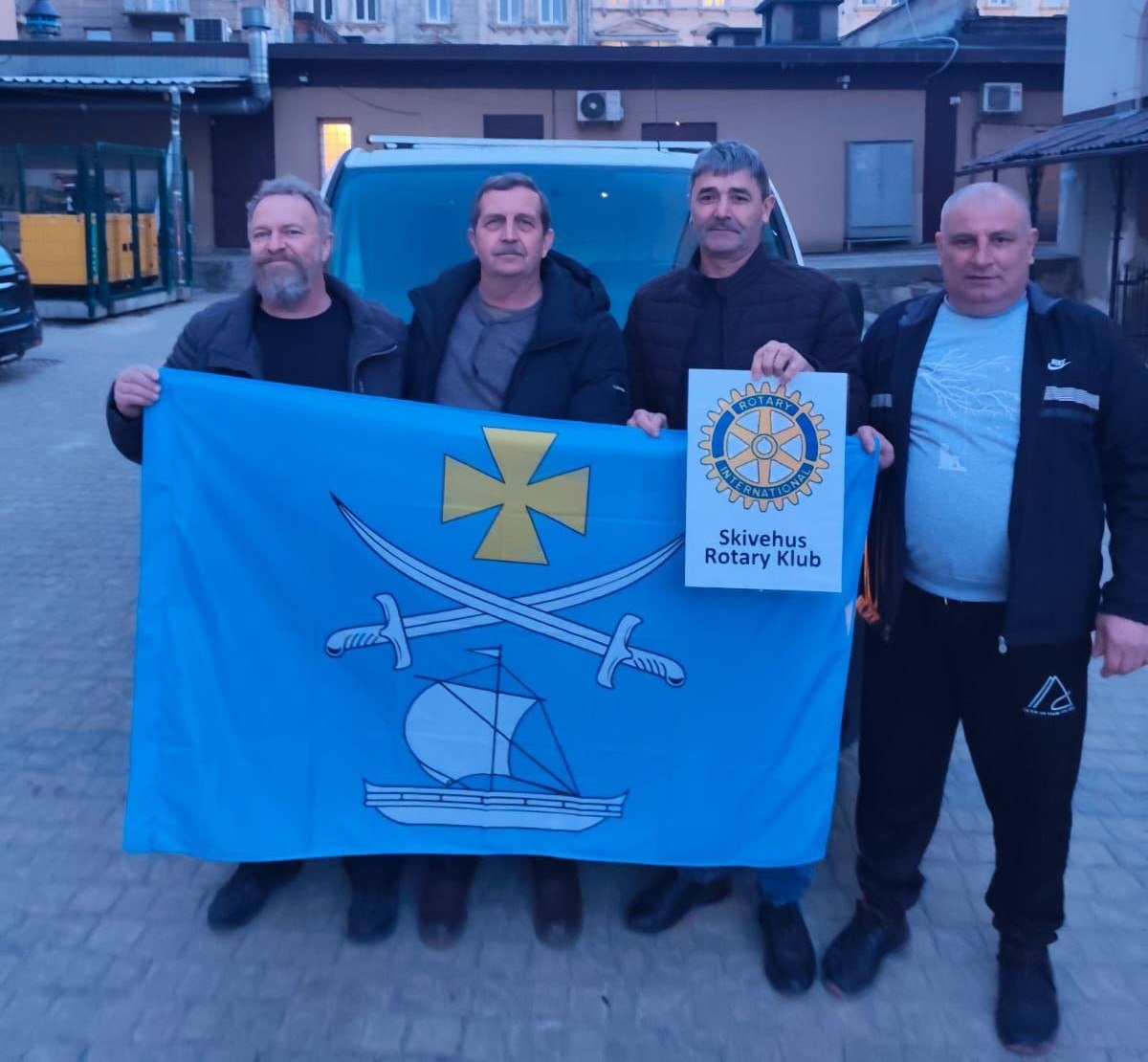 Четверо чоловіків стоять на вулиці перед автомобілем, тримаючи великий блакитний прапор із зображенням козацьких шабель, хреста та човна. Поруч вони тримають табличку з емблемою Rotary International та написом «Skivehus Rotary Klub».