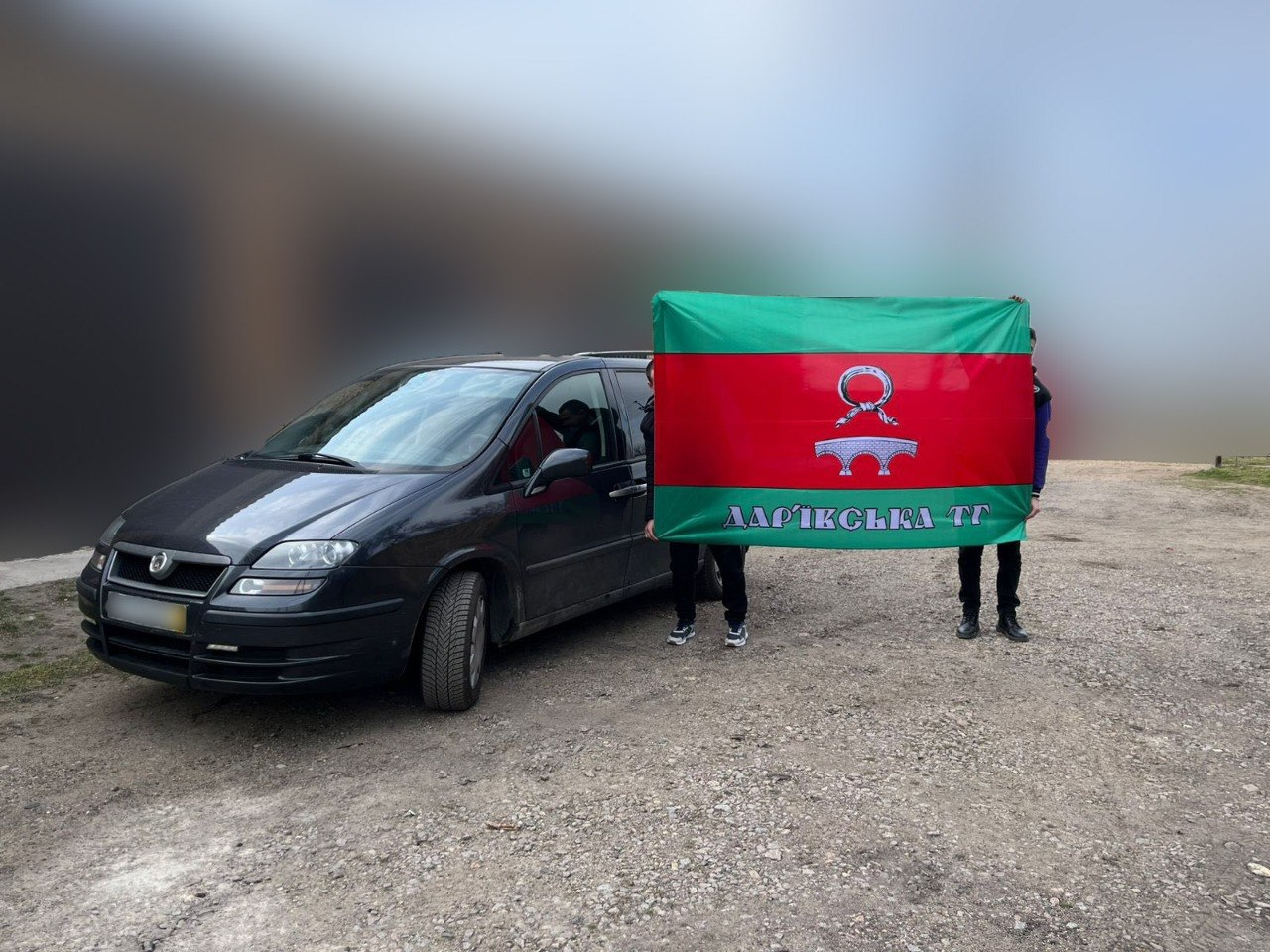 Двоє людей тримають великий червоно-зелений прапор із написом «Дар'ївська ТГ» та зображенням мосту. Вони стоять на ґрунтовій дорозі поруч із темним автомобілем, а фон зображення навмисно розмитий.